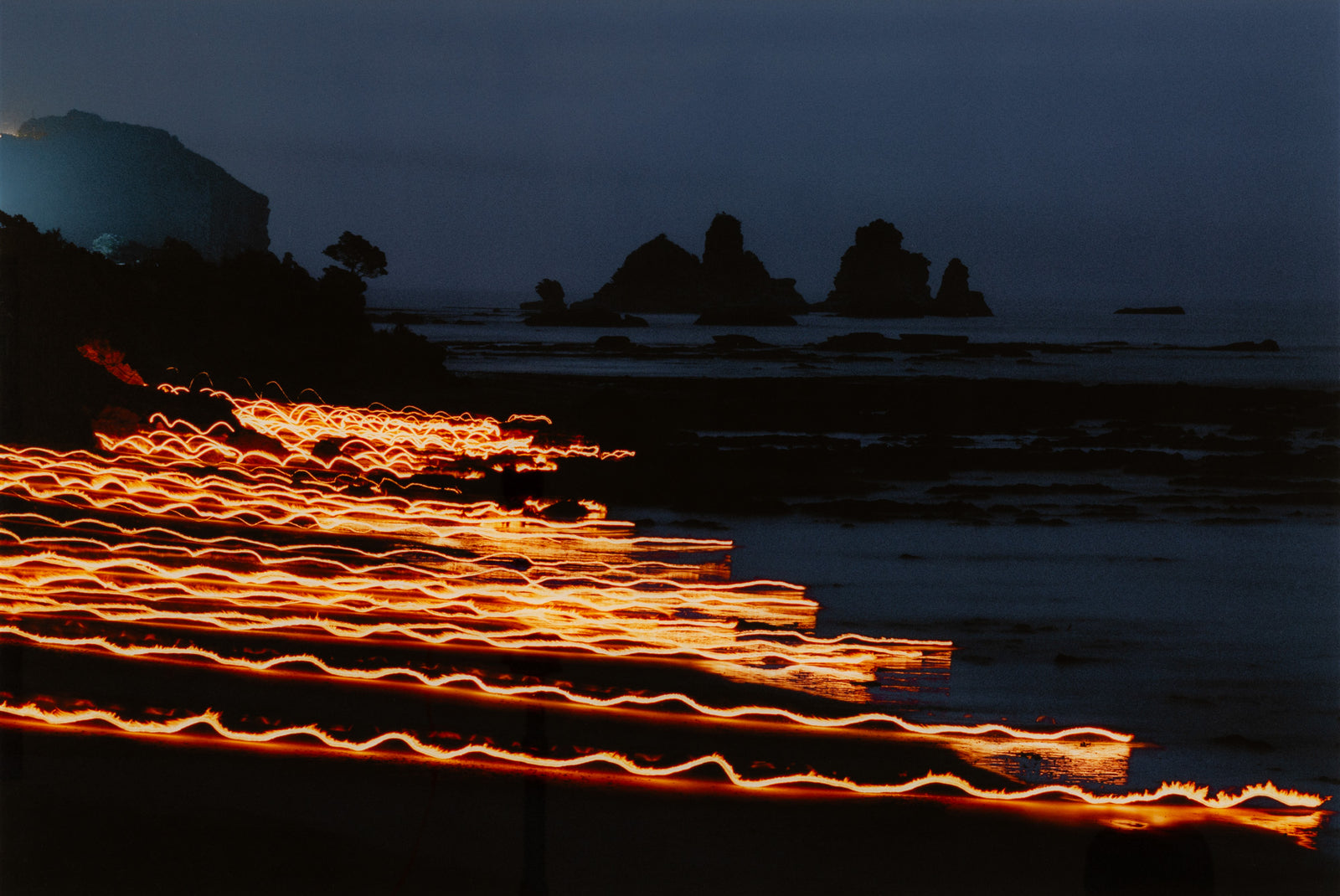 Photographie d’art d’André Wagner: Plusieurs lignes lumineuses dorées et ondulantes traversent en oblique un premier plan côtier sombre — Behind the lines, Neuseeland.