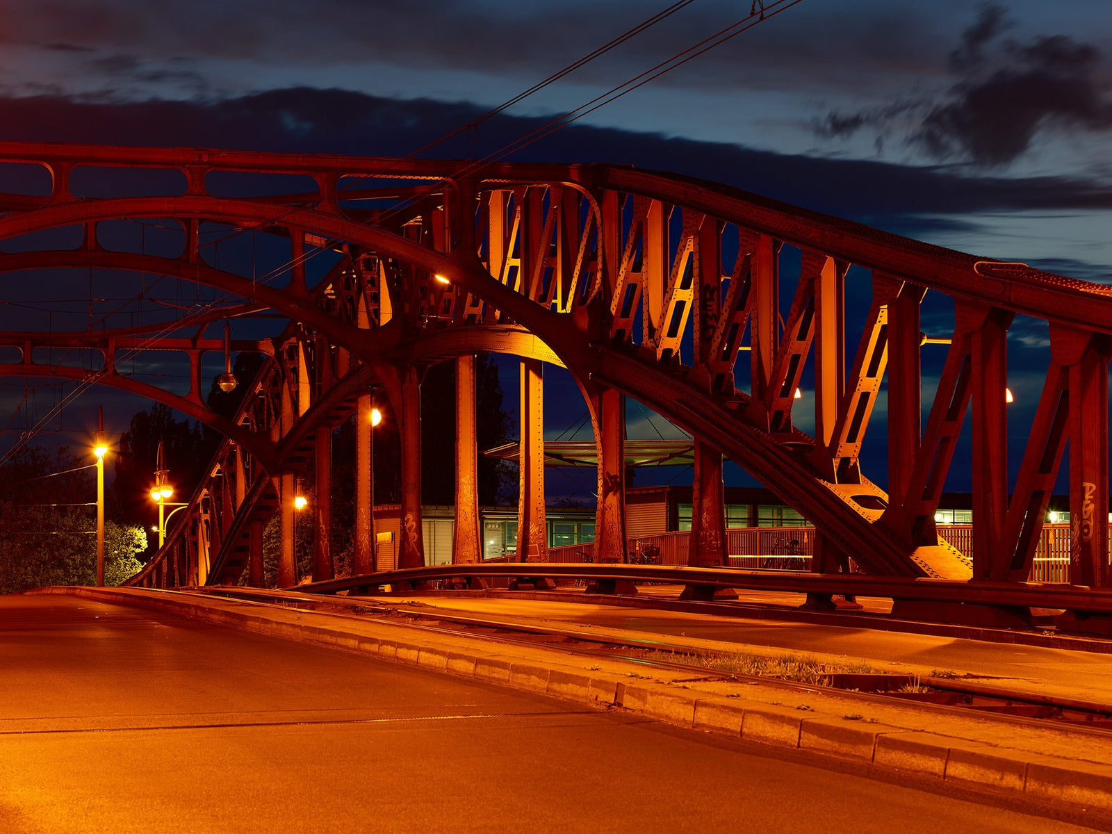 Fine art photography by André Wagner: The Bornholmer bridge carries its reddish illuminated steel arches over an empty roadway — Bornholmer brücke, Deutschland.