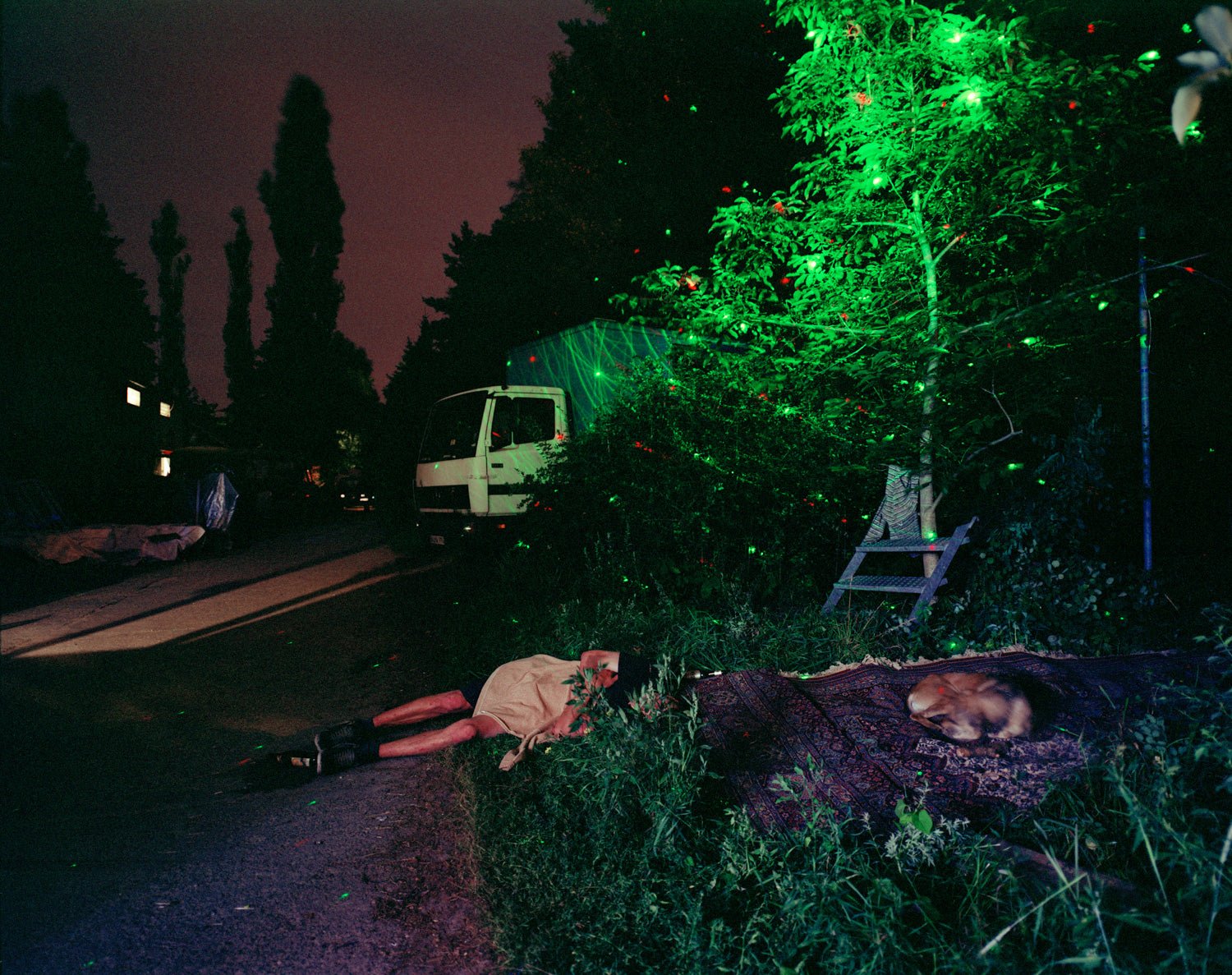 Fine art photography by André Wagner: A person lies beside a path; a dog rests on a patterned rug to the right — Chaos, Deutschland.