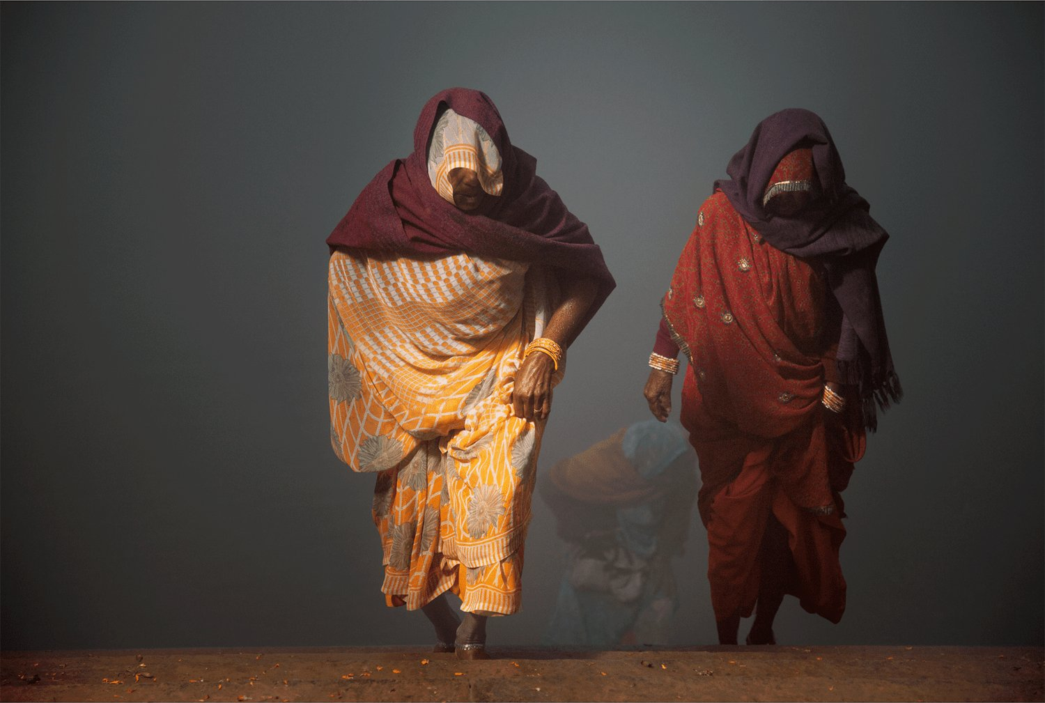 Fine art photography by André Wagner: Two women emerge clearly from grey mist, while another figure remains faint behind them — Coming back from yamuna river, Indien.