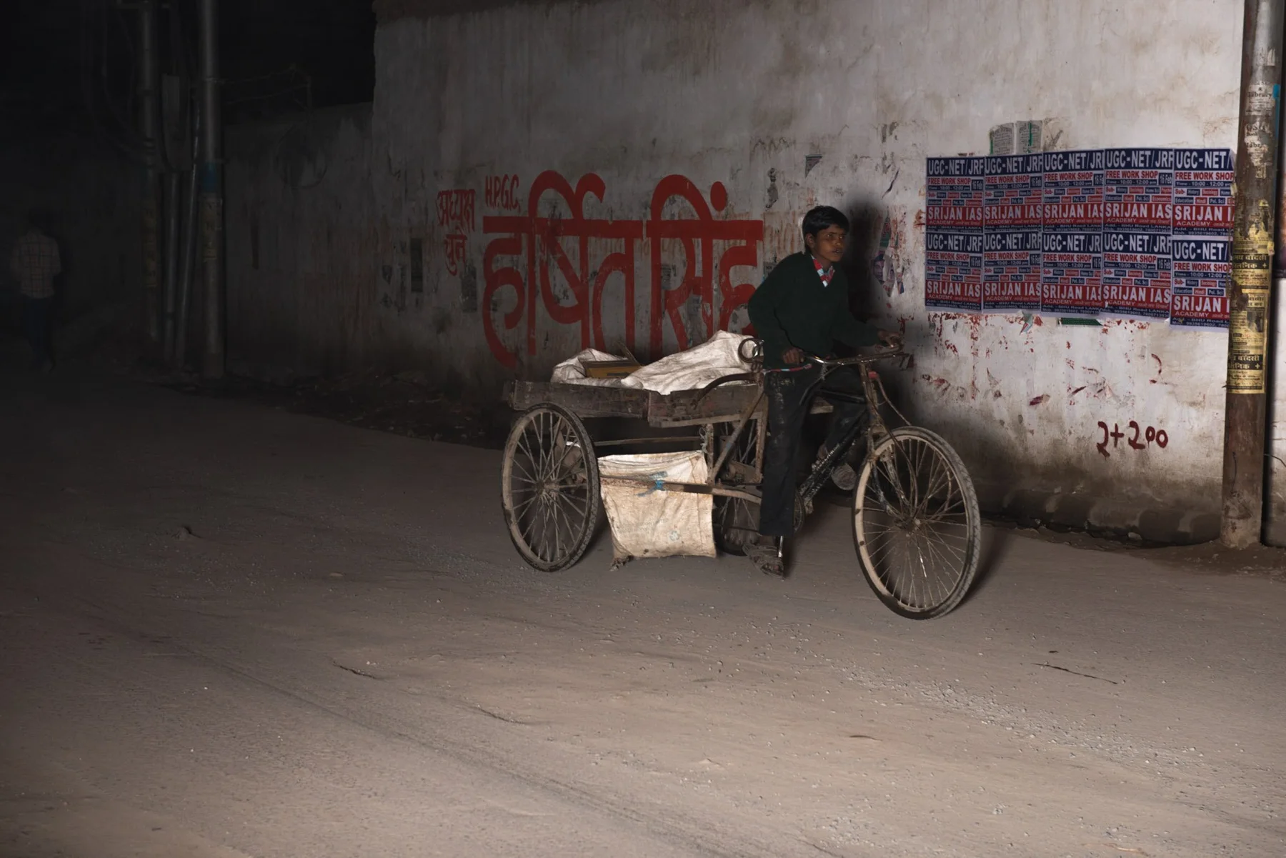A man stands at night with a bicycle and trucks in front of an inscribed wall.