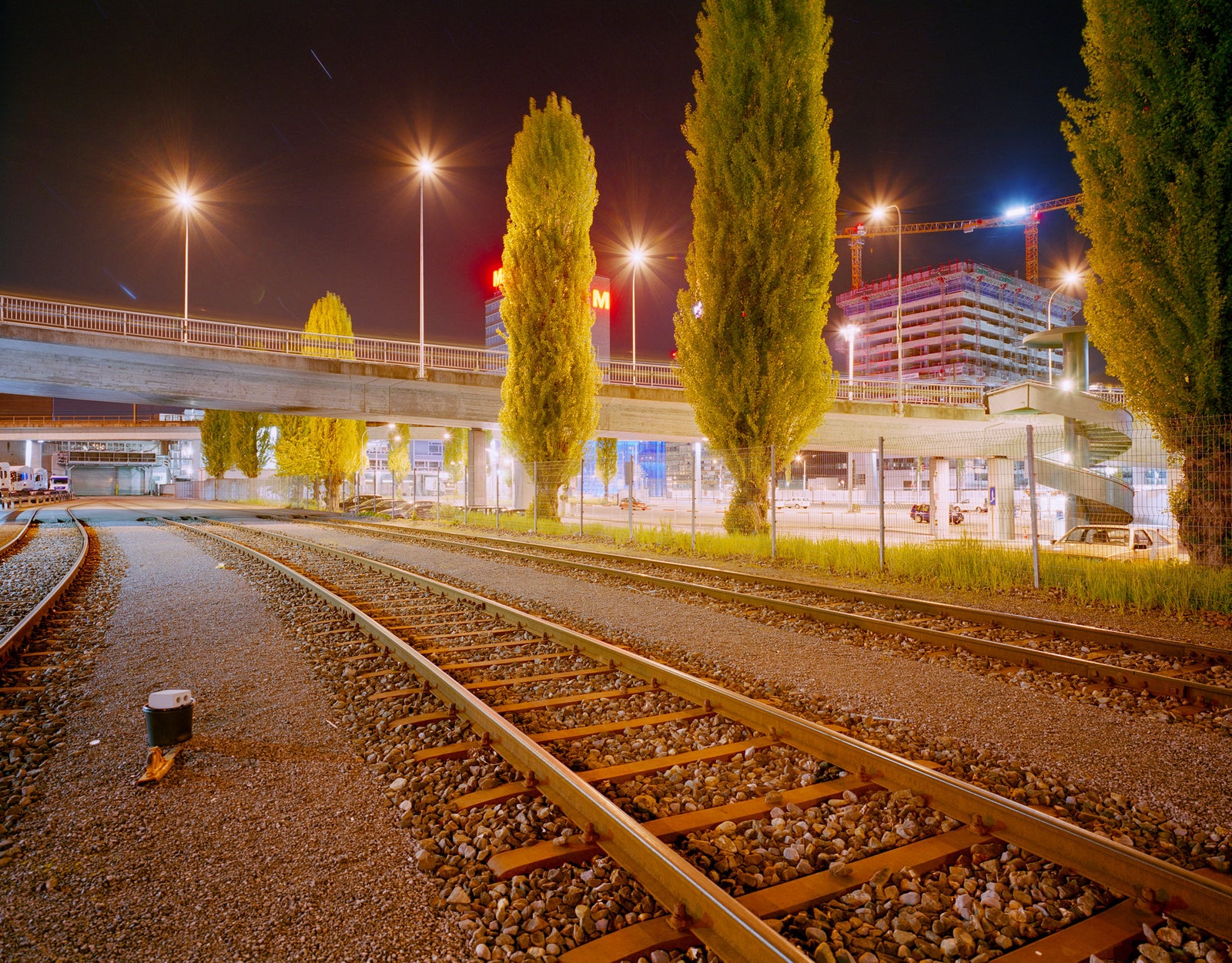 Fine art photography by André Wagner: Tracks curve between ballast and slender trees into an illuminated city — Five Stars.
