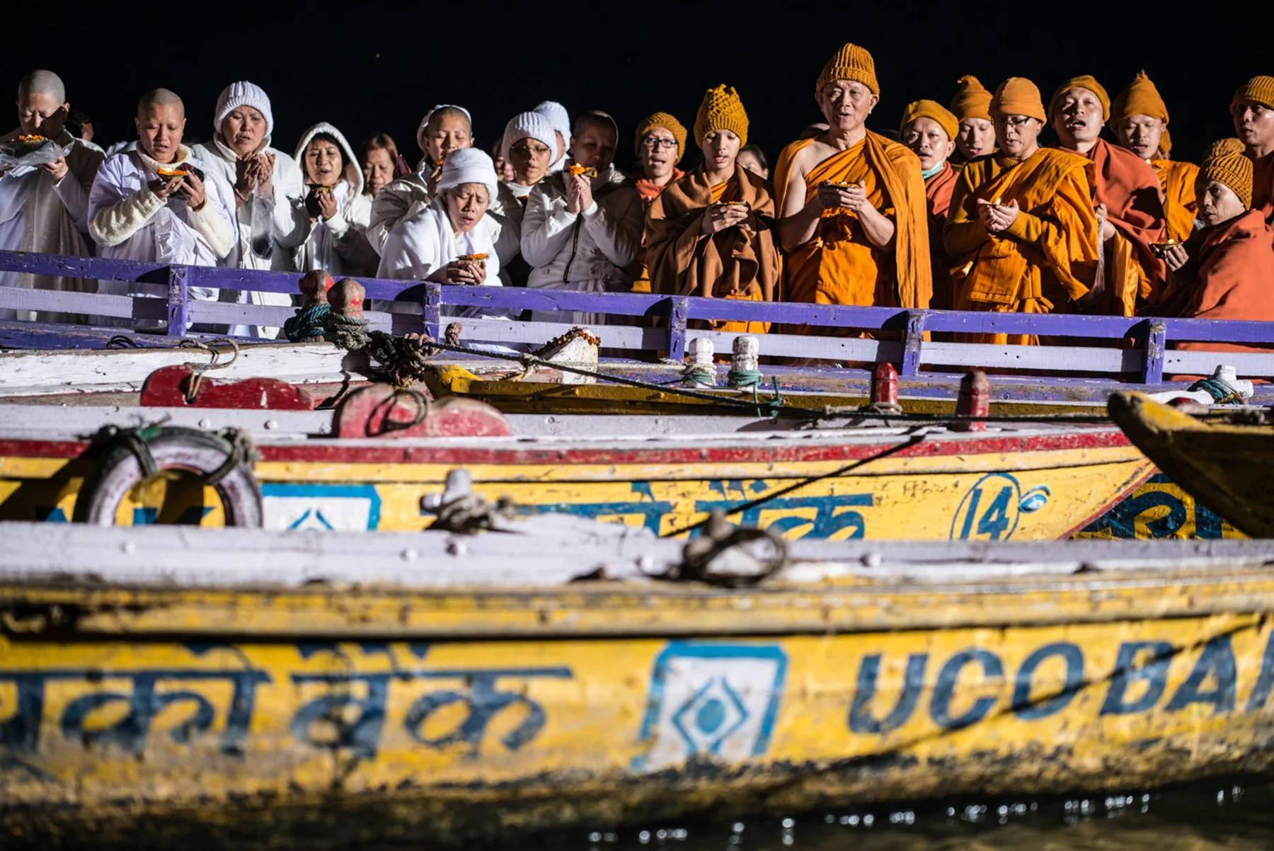 Group in bright and orange robes on a row of painted boats at night.
