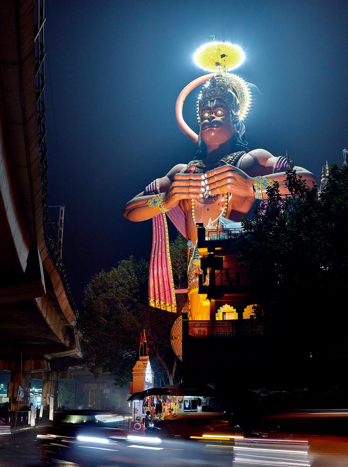 Fine art photography by André Wagner: A monumental Hanuman figure rises above a street at night — Hanuman temple in delhi, Indien.