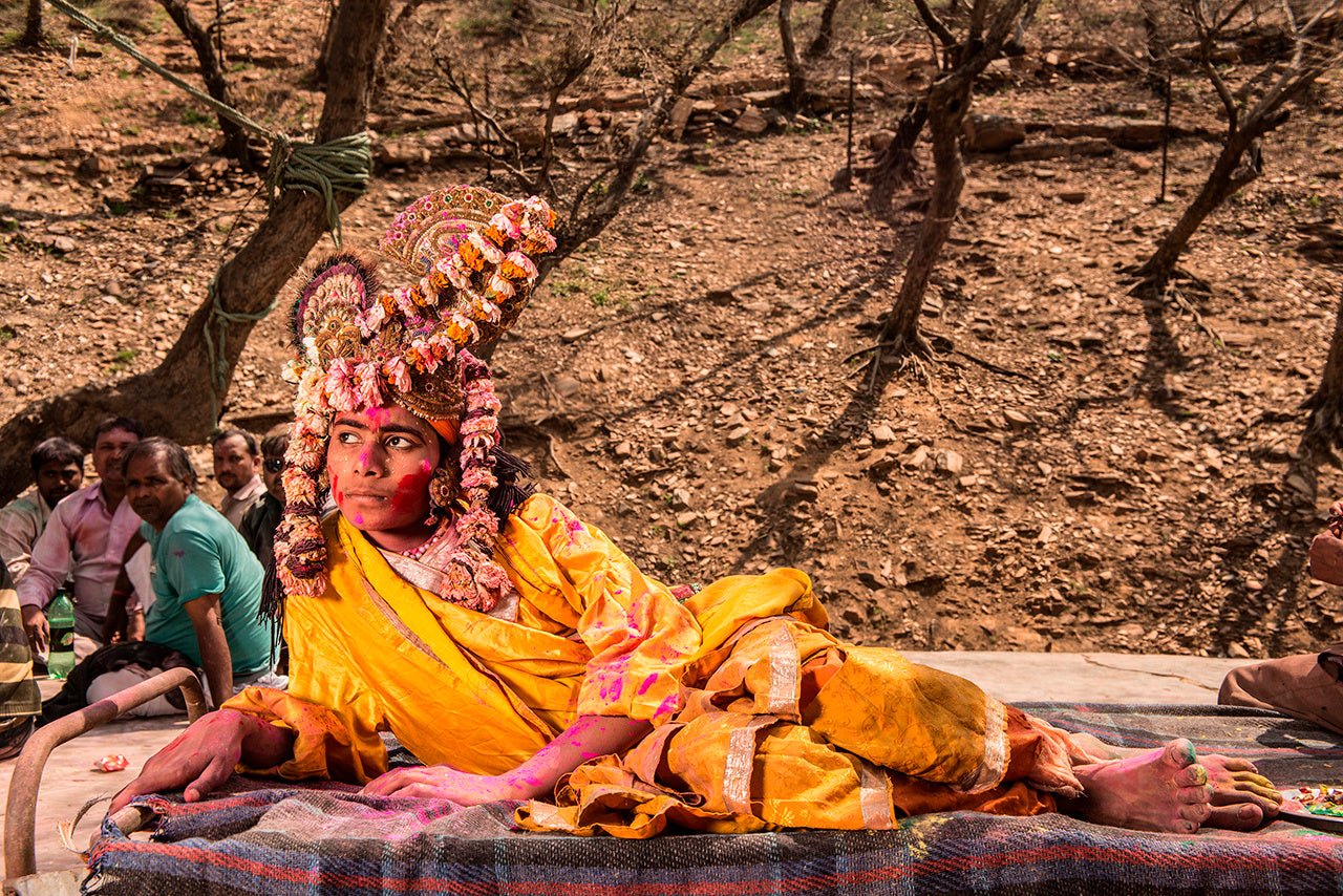 Photographie d’art d’André Wagner: Une personne vêtue de jaune est allongée sur le côté, sur une surface surélevée — Krishna plays holi, Indien.