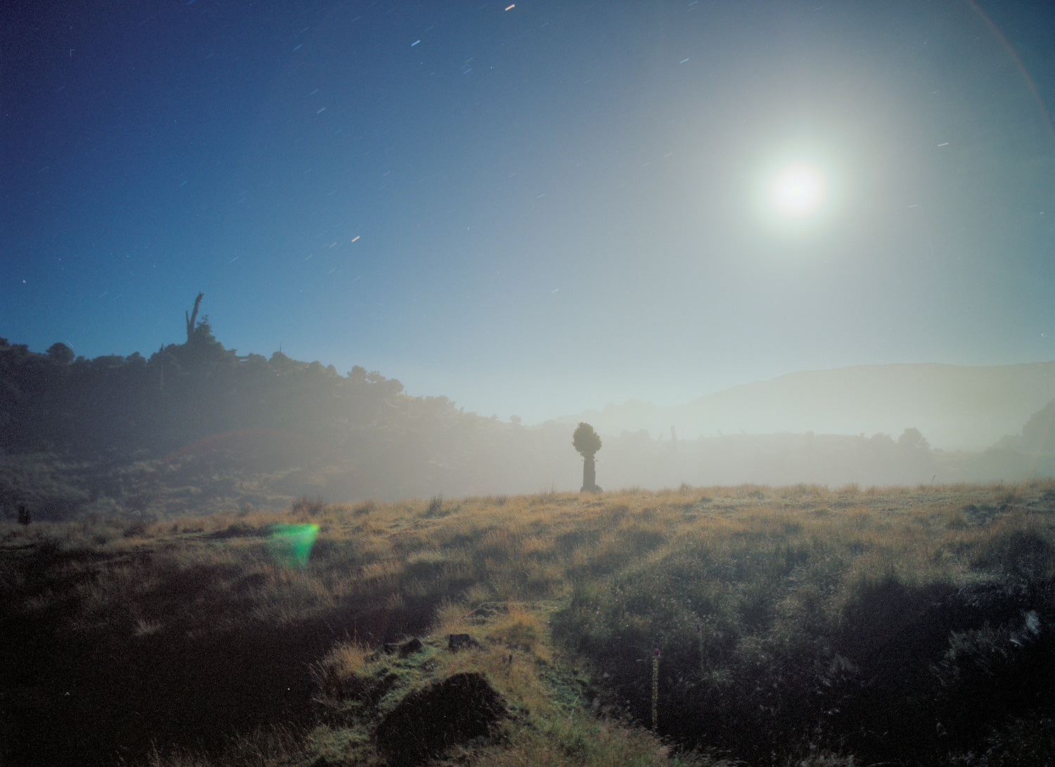 Fine art photography by André Wagner: A small human figure stands in an open low landscape beneath a bright point and pale glow in blue sky — Moonset.