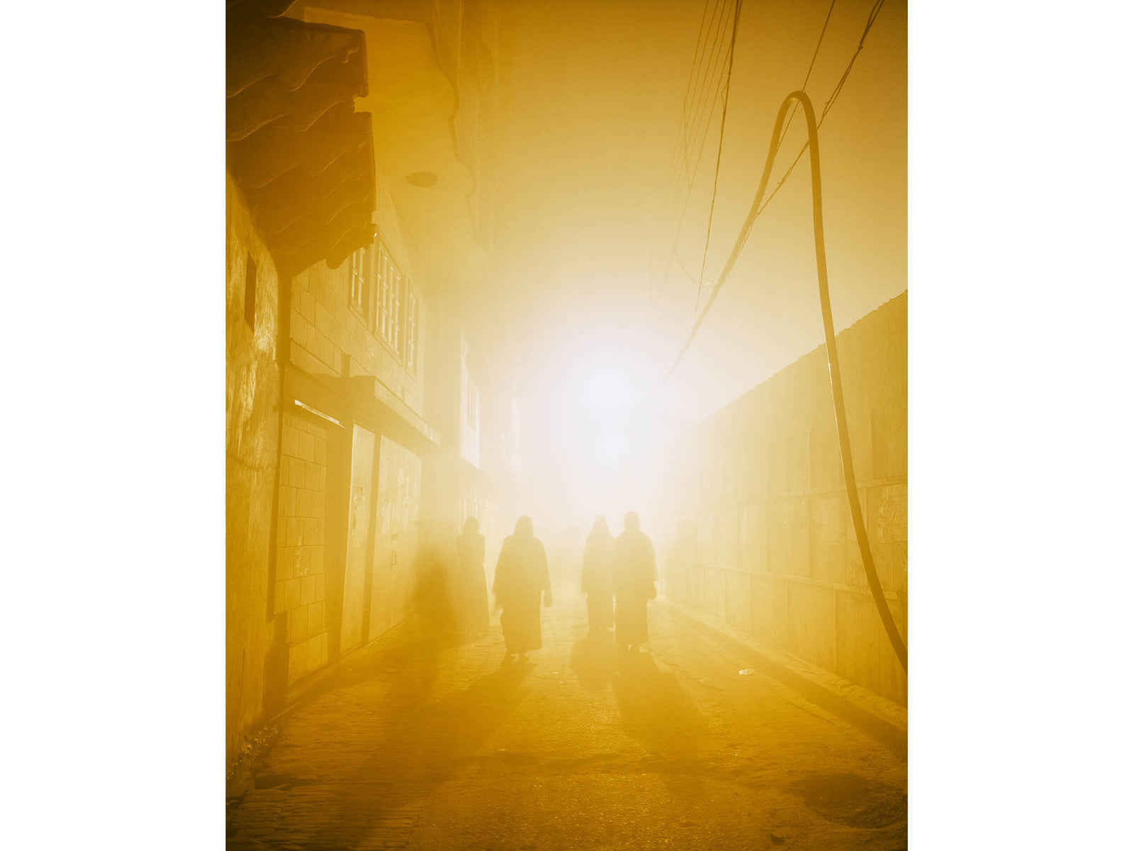 Fine art photography by André Wagner: A narrow lane is filled with yellow light and dense haze — Morning walk, Indien.