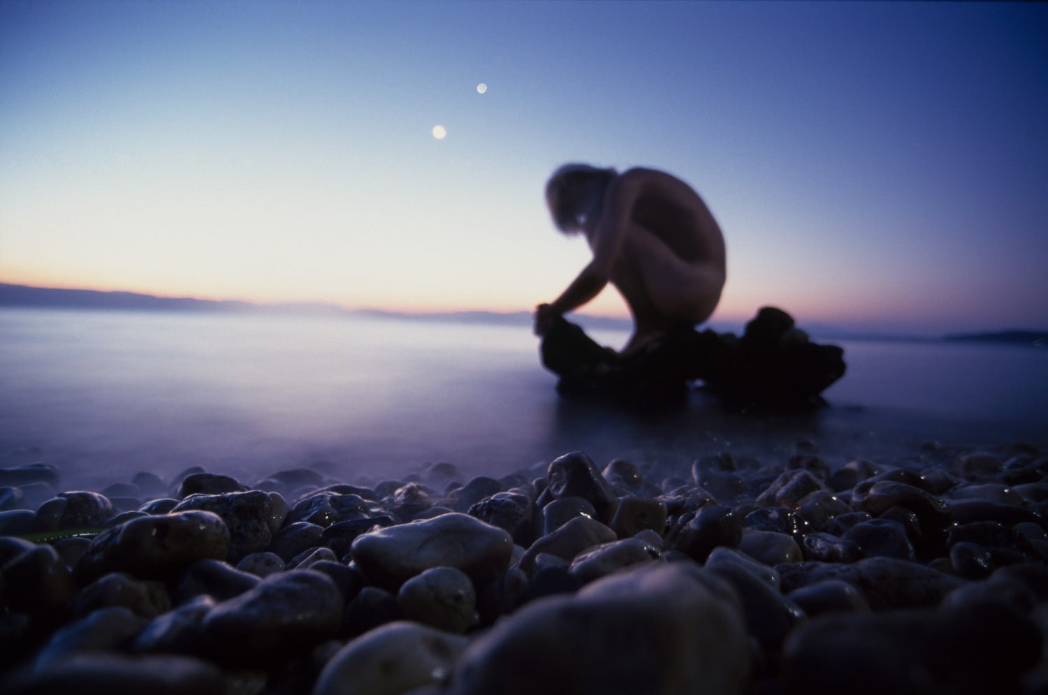Photographie d’art d’André Wagner: Une figure humaine nue est accroupie sur un rocher sombre, au bord de l’eau — New Born, Griechenland.