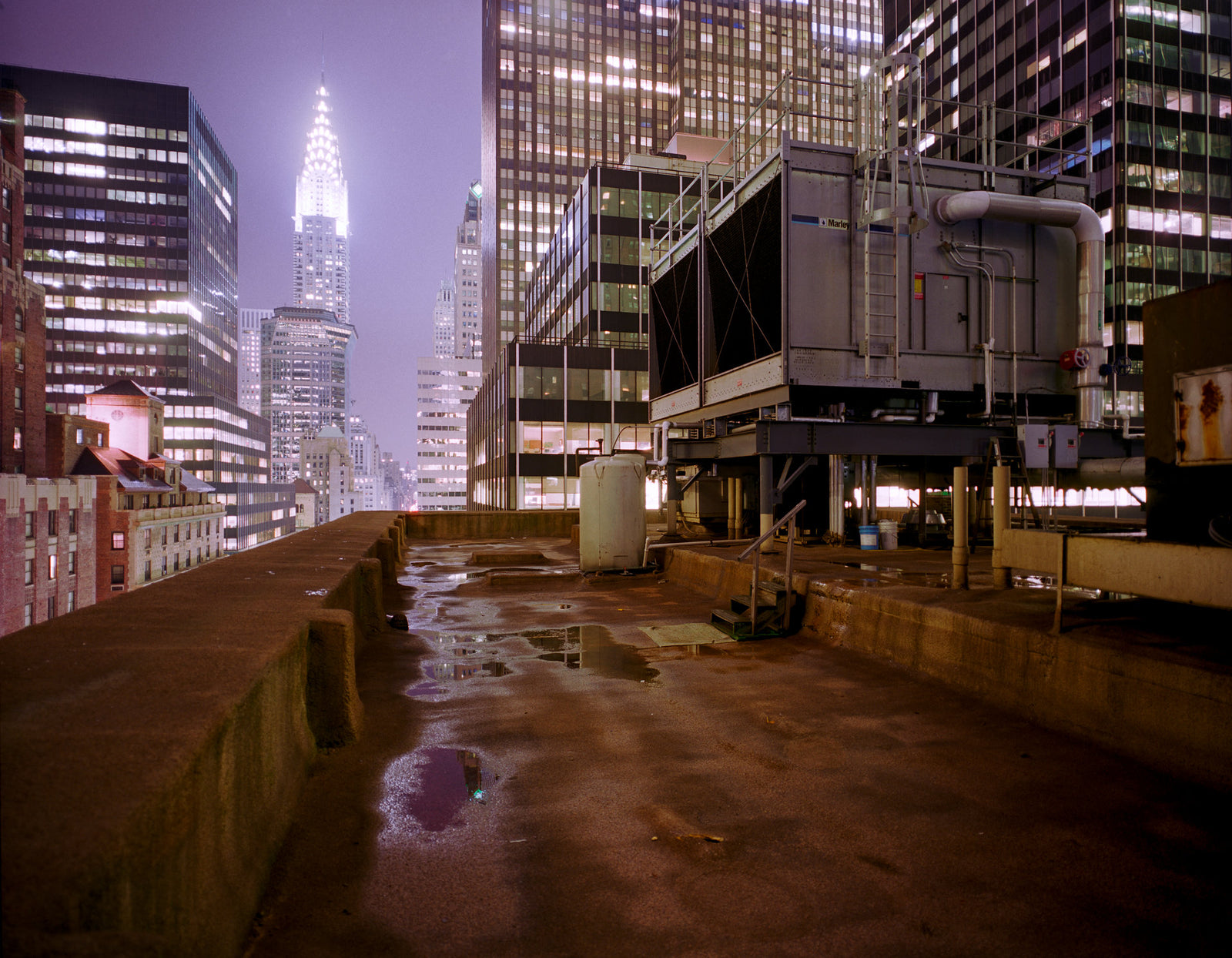 Photographie d’art d’André Wagner: Un toit mouillé conduit entre de hauts bâtiments vers une tour claire au loin — NY Roof 3.