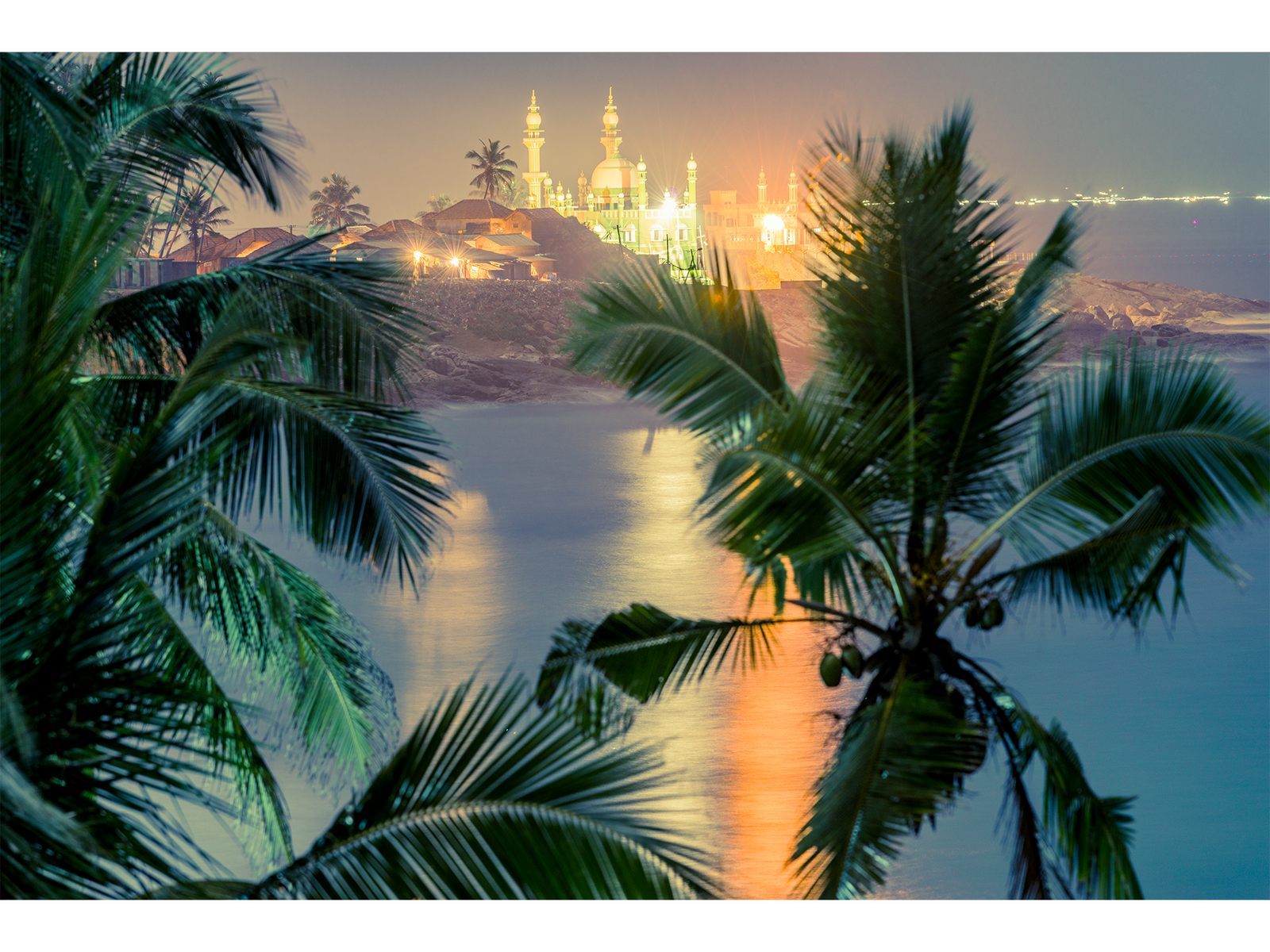 Fine art photography by André Wagner: Dark palm fronds frame a view across calm water — Ocean and mosque in kerala, Indien.