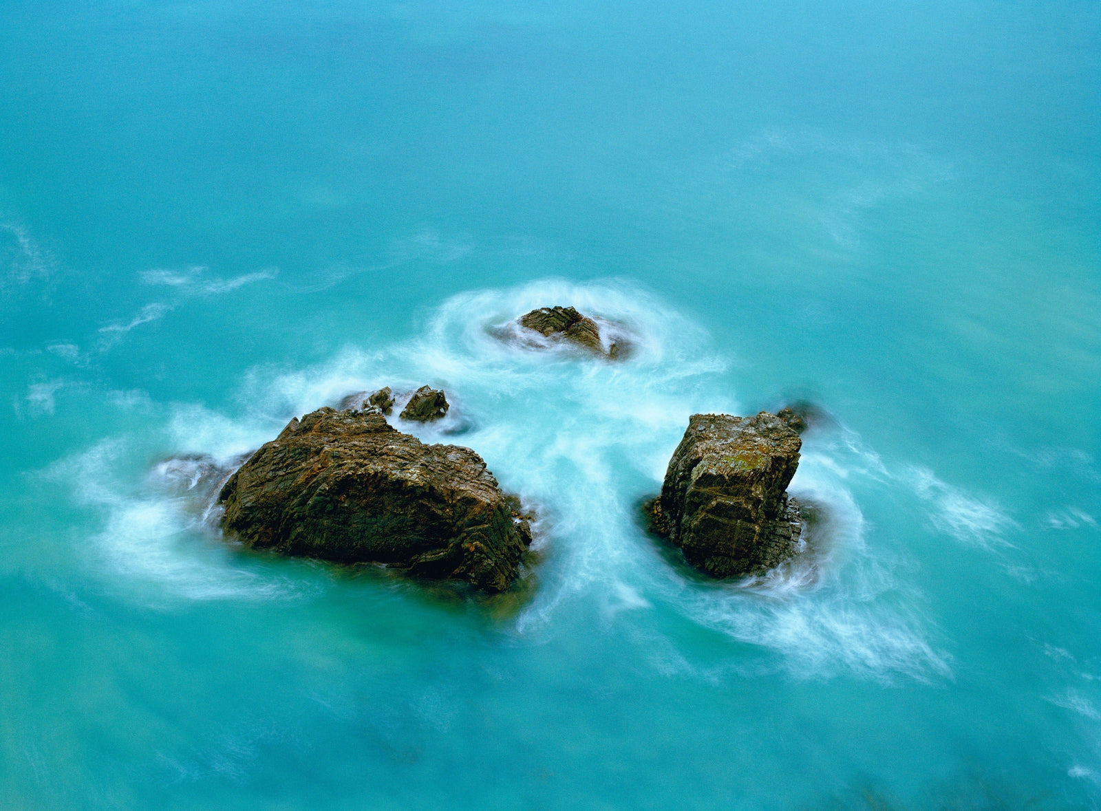Fine art photography by André Wagner: Several dark rocks lie within an almost frame-filling turquoise surface — Ocean, Neuseeland.