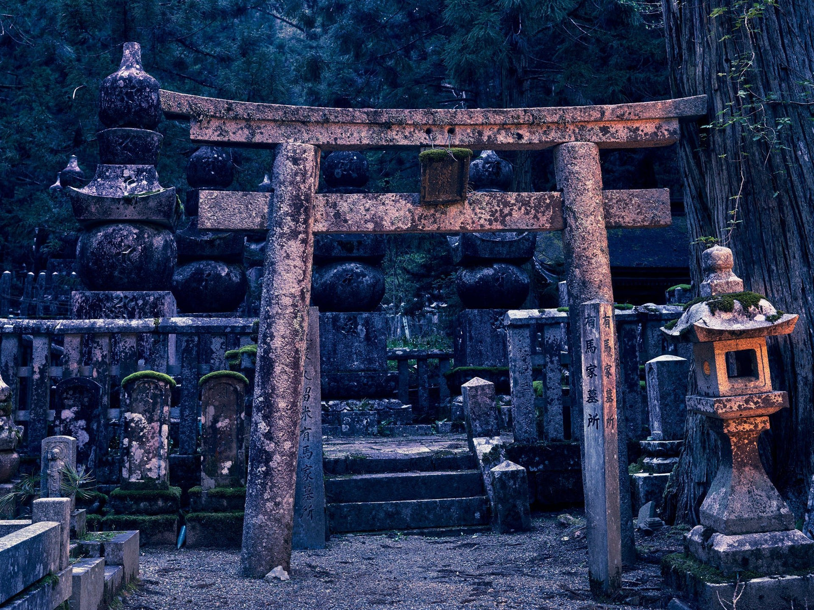 Photographie d’art d’André Wagner: Une porte de pierre se présente de face entre de grands arbres et de nombreuses formes de pierre plus petites — Oku-No-In Twillight, Japan.
