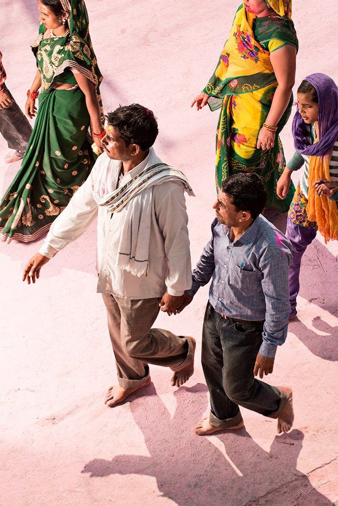 Photographie d’art d’André Wagner: Un homme vêtu de clair et une personne plus petite portant une chemise bleu plus sombre marchent côte à côte sur un sol rose — Pilgrim on Purple Dust 12, Indien.