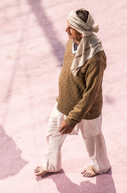 Fine art photography by André Wagner: A man in a pale headcloth, brown top and white trousers walks across largely open pink ground — Pilgrim on Purple Dust 4, Indien.