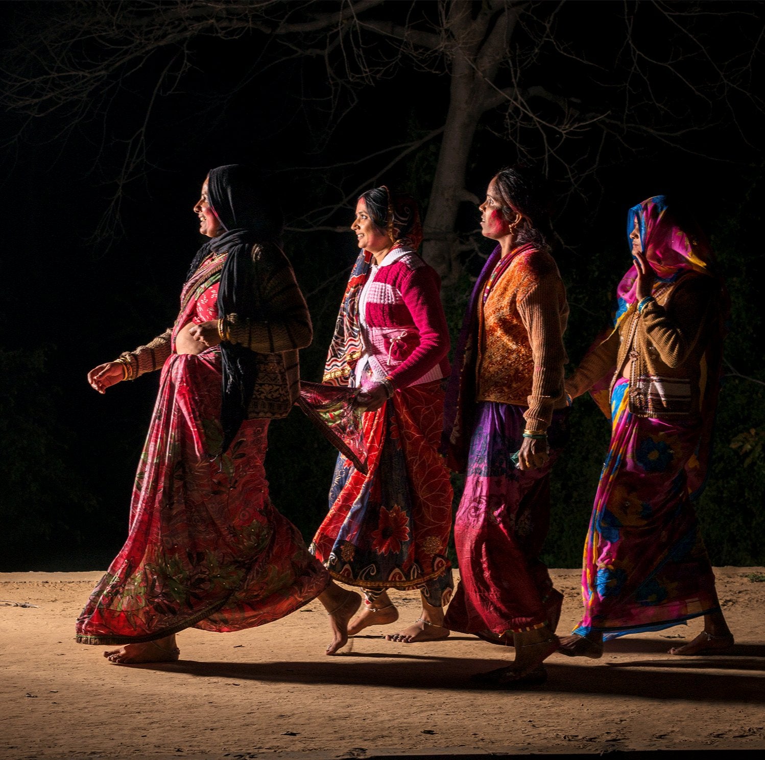 Fine art photography by André Wagner: Four people walk in a loose line before an almost black background — Pilgrims at govardhan 2, Indien.