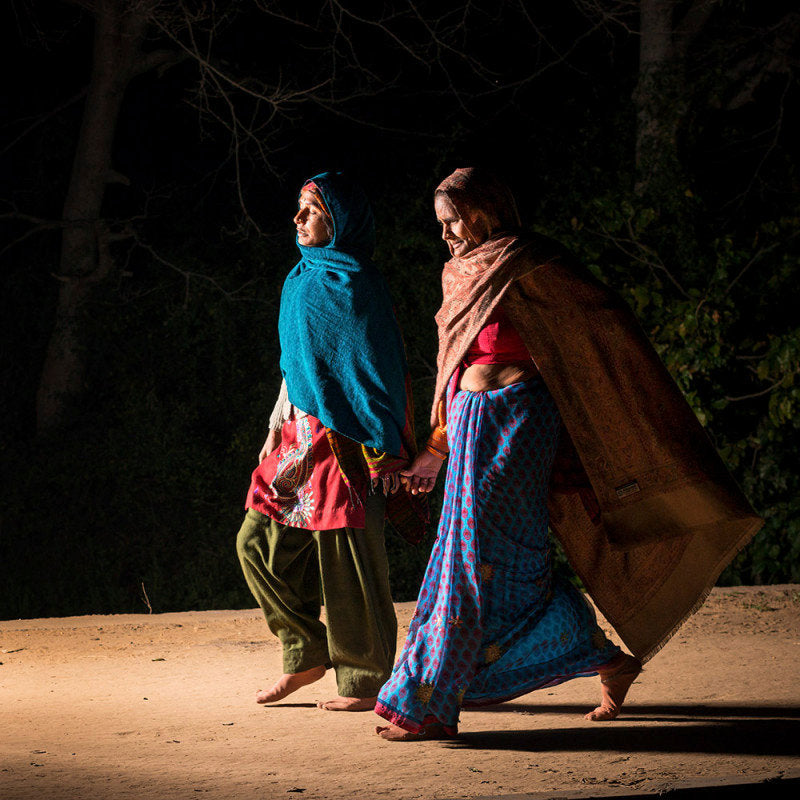 Fine art photography by André Wagner: Two women cross a lit strip before dark trees — Pilgrims at Govardhan 9, Indien.