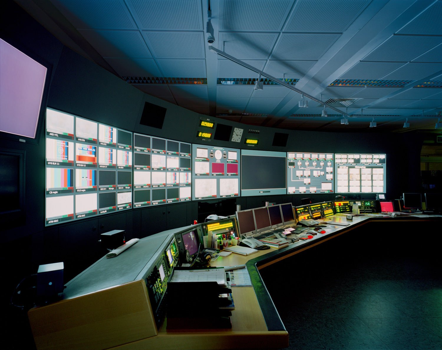 Fine art photography by André Wagner: A curved wall of small screens extends through a dark control room — Programs, Deutschland.