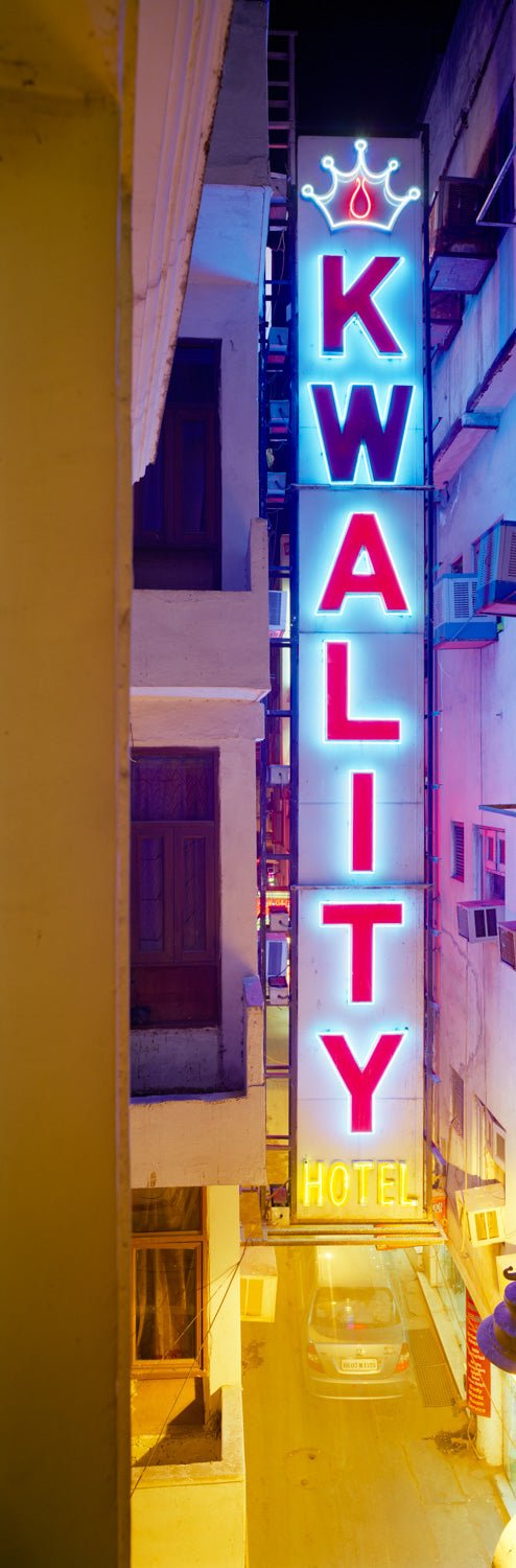 Photographie d’art d’André Wagner: Une enseigne lumineuse étroite et verticale porte des lettres colorées devant une façade sombre — Quality, Indien.