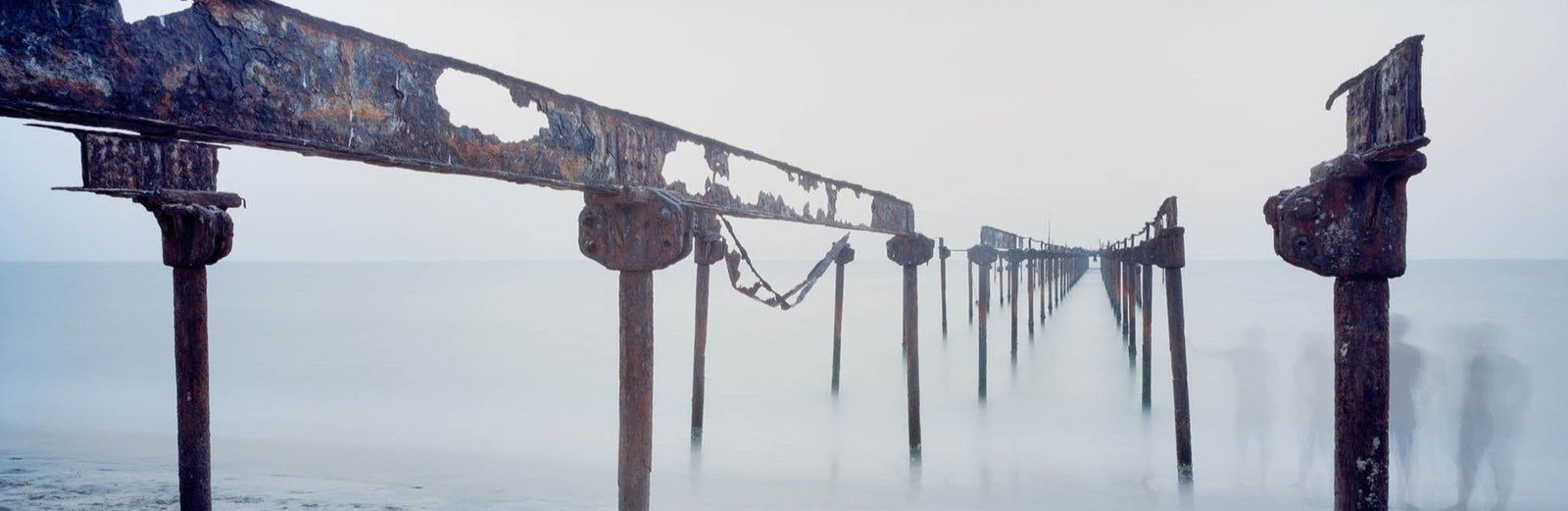 Photographie d’art d’André Wagner: Les poutres rouillées s’étirent au-dessus de l’eau, du bord proche de l’image jusqu’à un point de fuite lointain — Rust bridge, Indien.