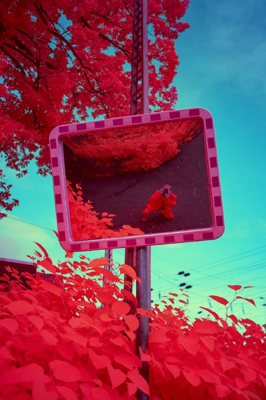 Photographie d’art infrarouge d’André Wagner: Un miroir routier apparaît de biais dans l’image et contient une seconde vue : entre un feuillage rouge et un sol sombre se tient une personne qui photographie, l’appareil devant le visage — Signal of the Self.