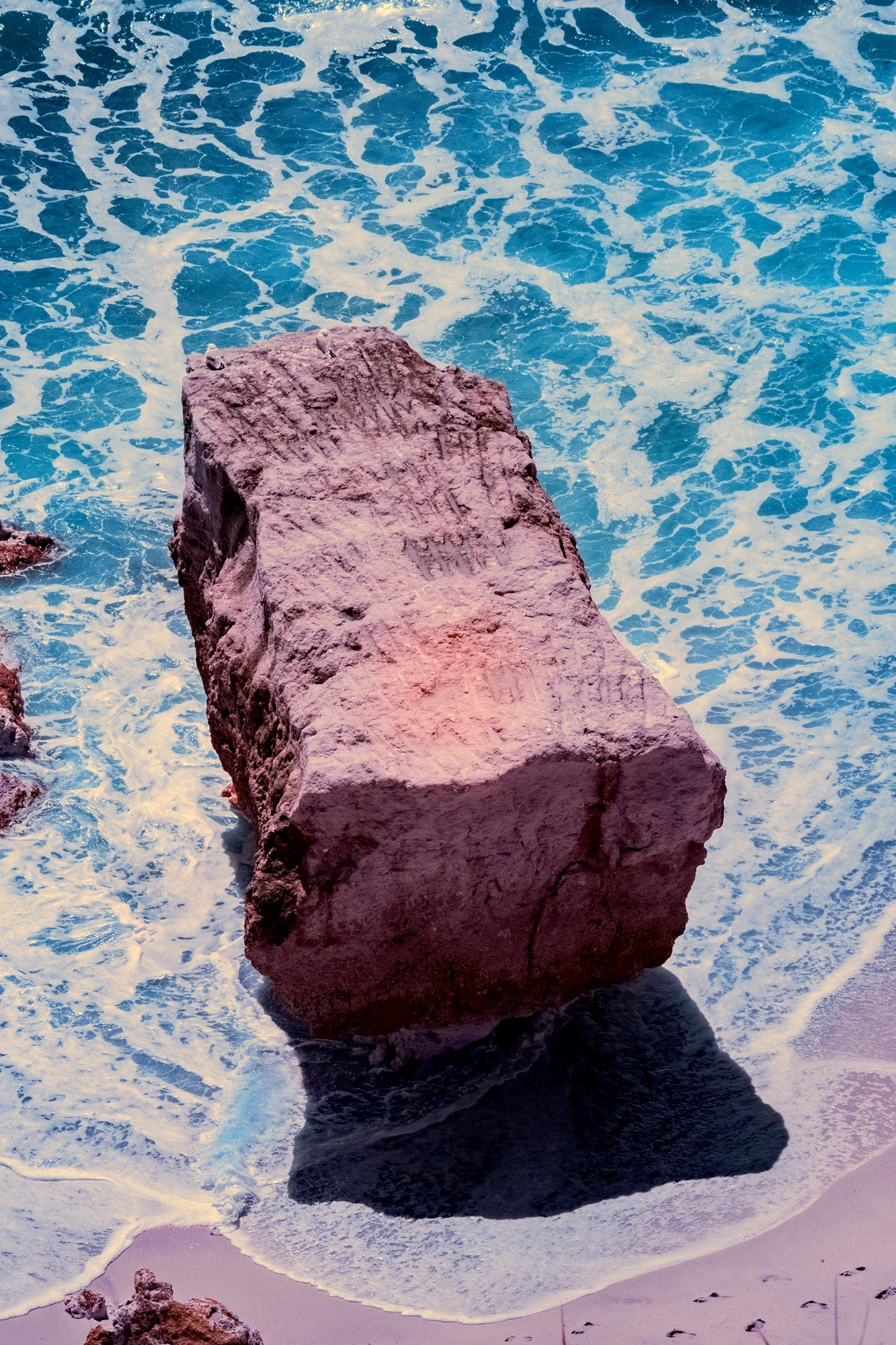 Photographie d’art infrarouge d’André Wagner: Un rocher anguleux repose comme un corps pesant dans l’eau turquoise — The Last Rock.