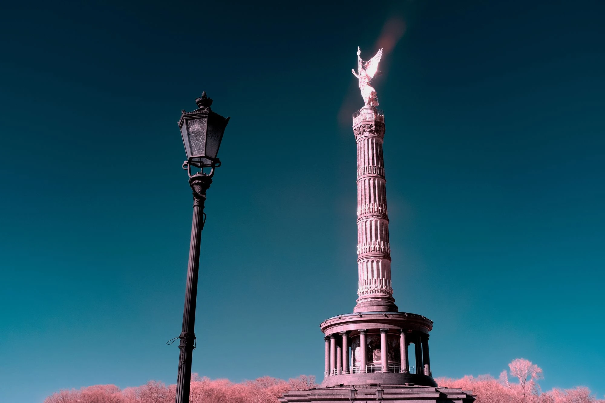 Photographie d’art infrarouge d’André Wagner: Un lampadaire et la colonne de la Victoire s’élèvent côte à côte dans le ciel bleu sombre — Through Time’s Lens: Infrared Imagining of Contemporary Struggles.
