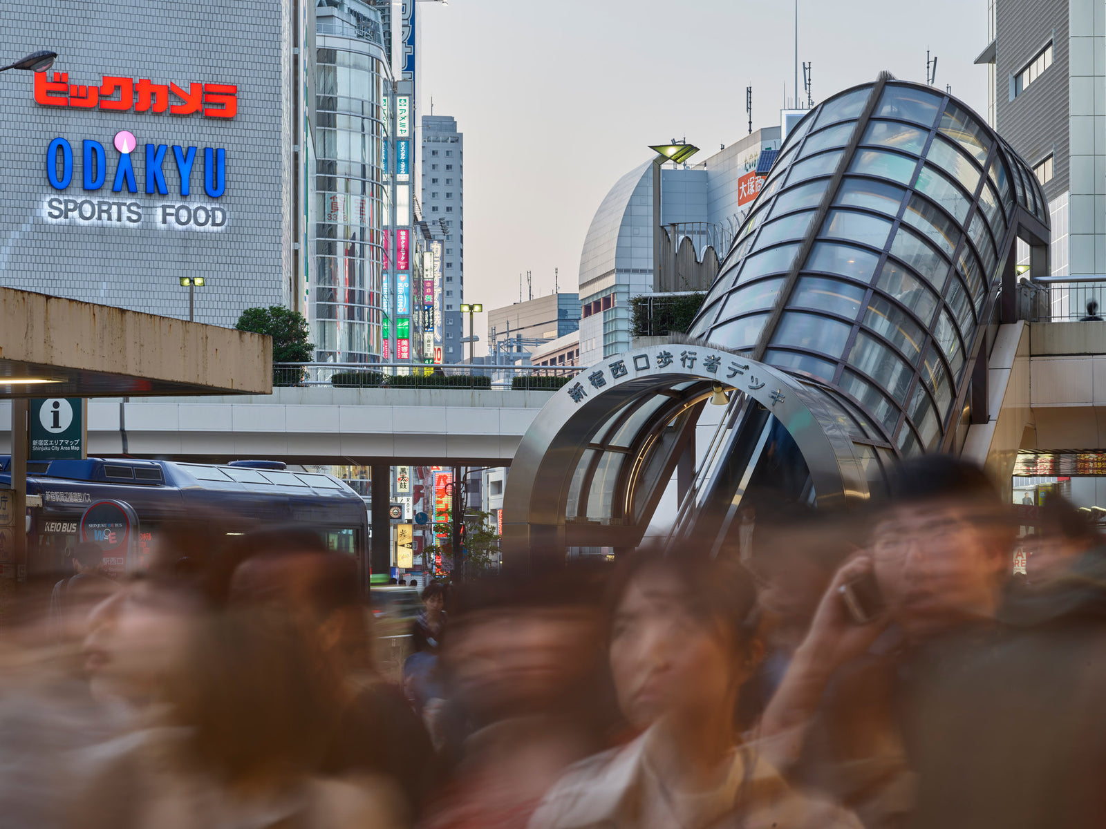 Photographie d’art d’André Wagner: Un accès vitré et courbe se dresse en oblique devant une grande façade commerciale — Tokyo Shinjuku 7.