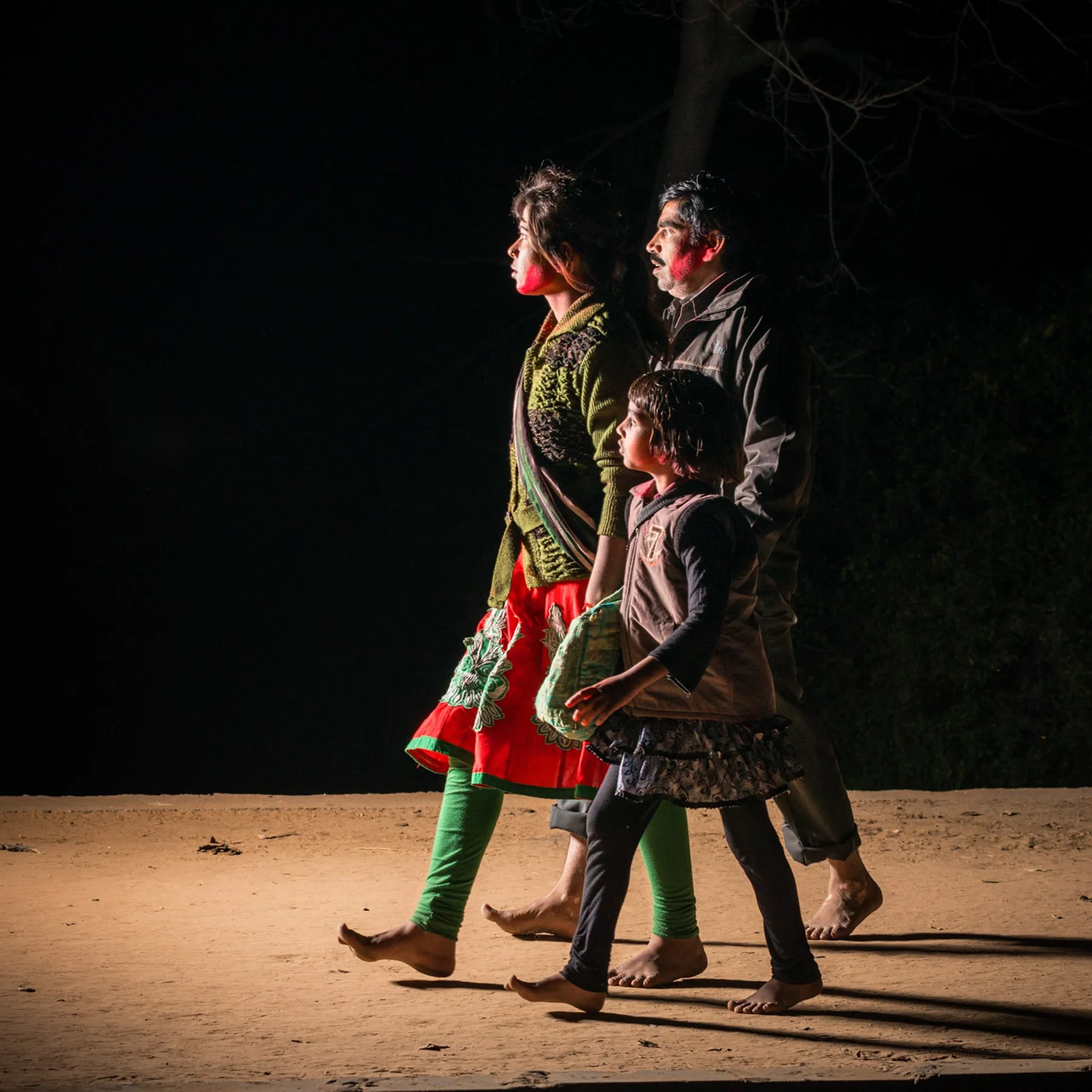 Two women and a child walk across a lighted street at night.