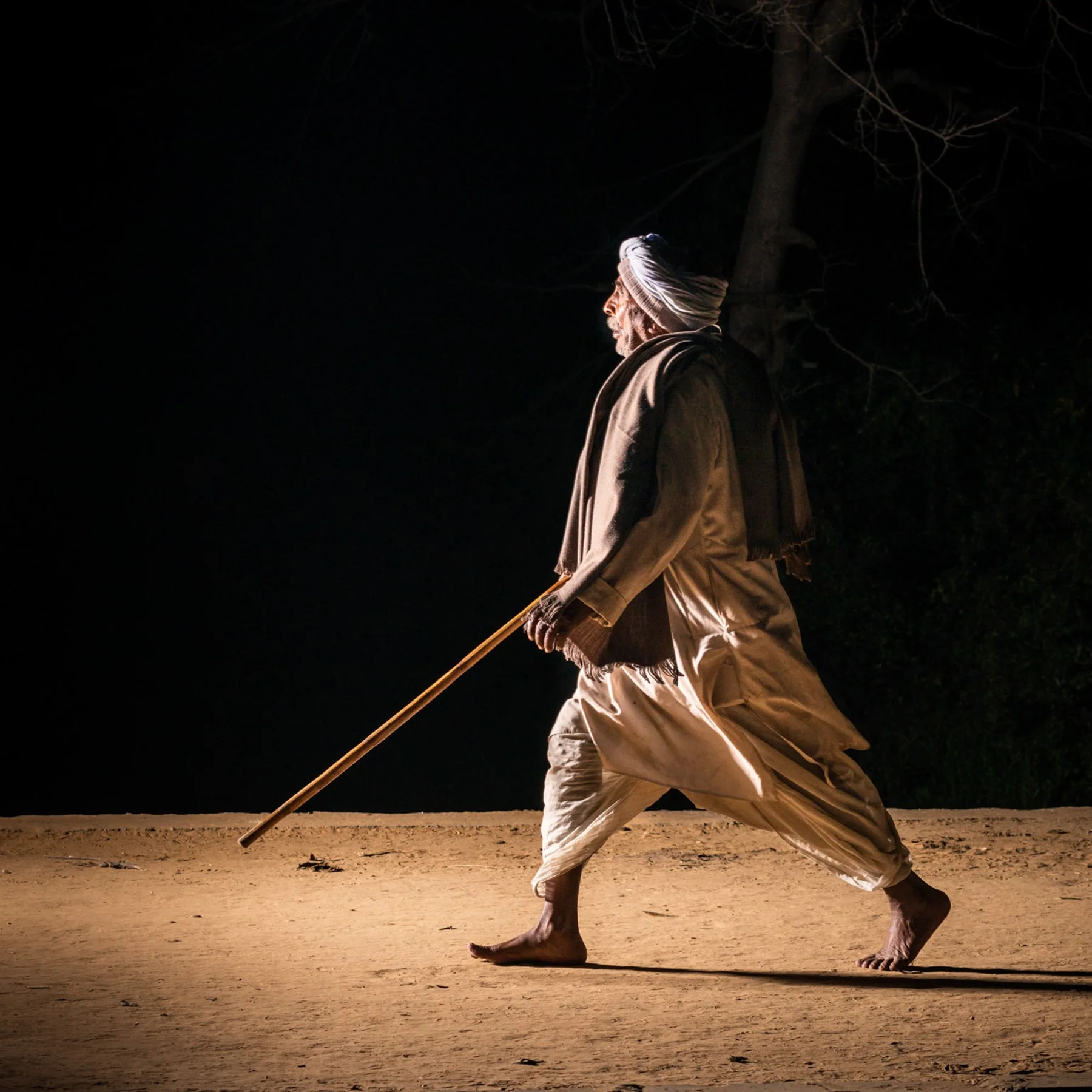 Un homme en turban portant un long bâton dans une rue éclairée la nuit.