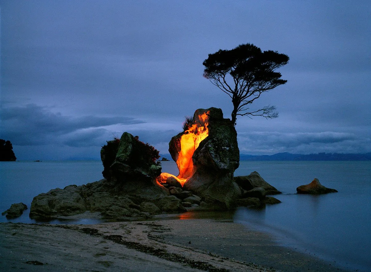 A rock in the water with a burning inner fire, a tree on the rock, dark skies and water in the background.