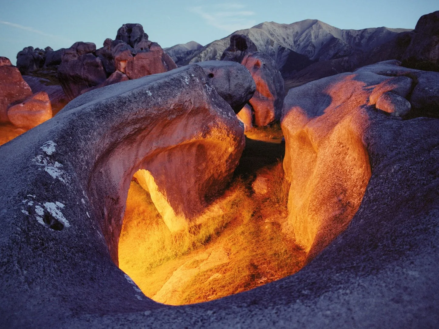 A lava flow in a volcanic landscape at sunset, surrounded by large rocks and mountains.