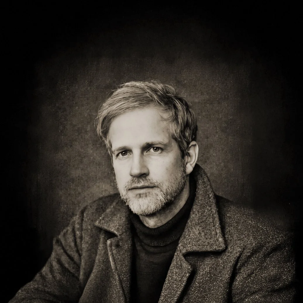 Portrait en noir et blanc d&rsquo;un homme aux cheveux mi-longs, barbu, &agrave; l&rsquo;expression s&eacute;rieuse, portant un pull et un manteau sombres sur fond sombre.