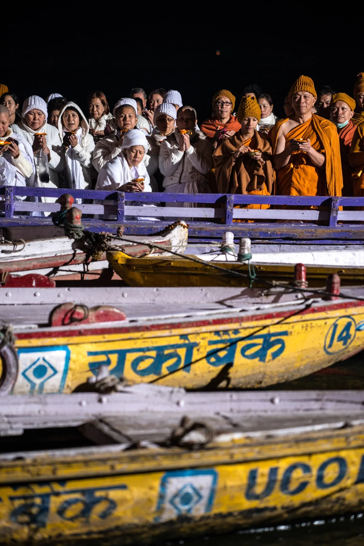 Des personnes en vêtements blancs et orange assises la nuit derrière des bateaux peints de couleurs vives.