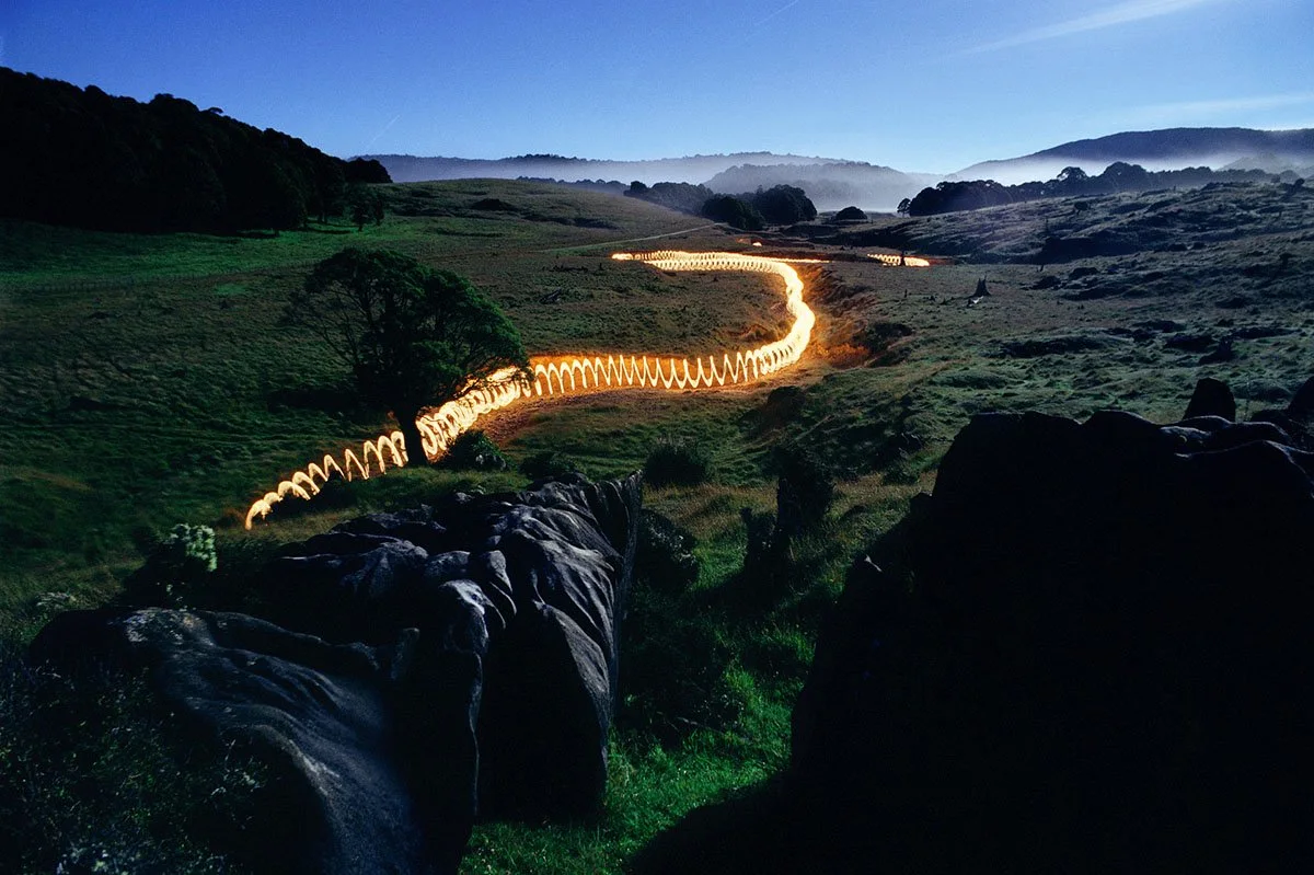 Landscape at dusk with hills, trees and an illuminated path that winds through the valley.