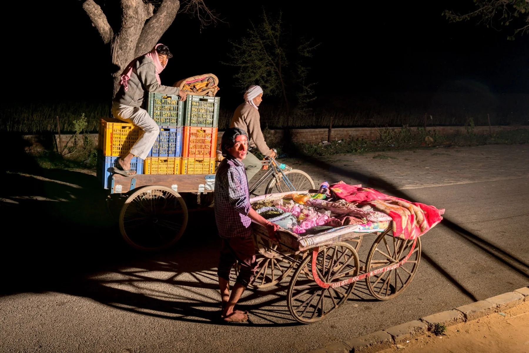 Des personnes près de chariots de vente éclairés sous un arbre, dans une rue nocturne.